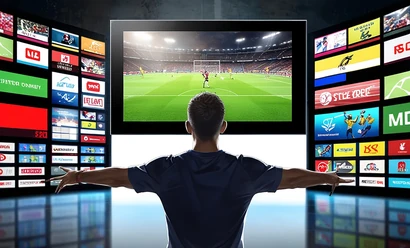 Kanal logolarıyla çevrili bir kişi, merkezdeki büyük ekranda futbol yayınını izliyor